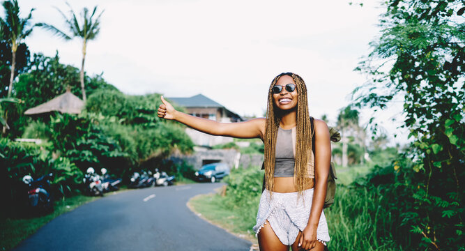 Cheerful African American hitchhiker with hand up catching automobile transport at roadside for getting to travel destination during wildlife style of trip, joyful female tourist enjoying vacations