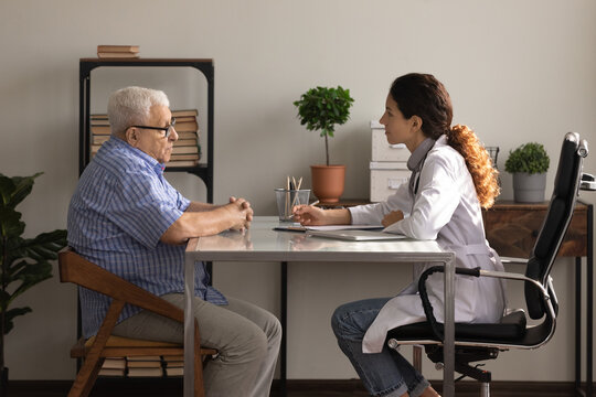 What Is Your Complaint. Side View Of Older Man Patient Meeting Young Woman Attending Physician For Regular Medical Checkup. Senior Male Clinic Visitor Talking With Latina Female Doctor At Appointment