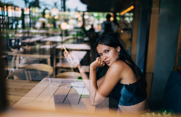 Pensive young ethnic female blogger in cafe with smartphone and netbook