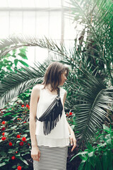 Attractive young woman stands among tropical plants. Woman in white blouse and black leather fringe...