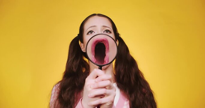 Young funny attractive caucasian girl with long braids holding and play magnifying glass in front of camera and makes faces and grimaces on red backgorund. Comic humor theme with magnifying face
