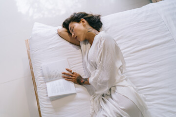 Thoughtful woman with book sleeping on bed