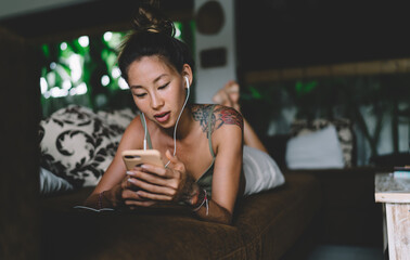 Ethnic woman in earphones using smartphone lying on sofa