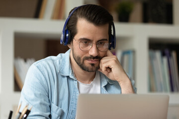 Close up focused man in glasses and headphones using laptop, looking at computer screen, watching webinar or training, studying online, student listening to lecture, involved in internet lesson