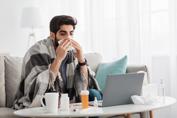 Ill arab man covered in warm blanket looking at laptop screen and sneezing nose in paper tissue, sitting on sofa