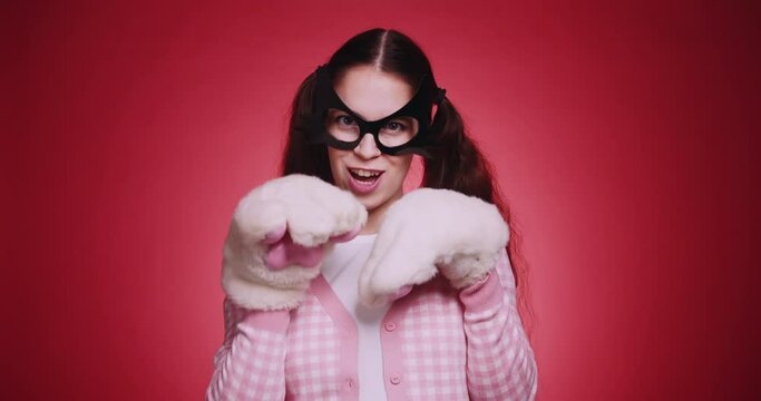 Cute girl in a cat mask and pink clothes with plush fluffy paws flirts with cat gestures in front of the camera on red background. Catwoman funny cosplay masquerade