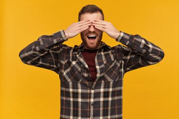 Cheerful male, handsome guy with brunette hair and beard. Wearing checkered shirt and accessories. Cover eyes with palms and broadly smiling. Hide and seek. Stand isolated over yellow background