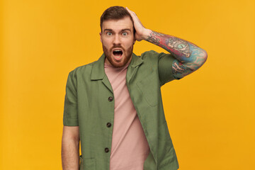 Bewildered male, shocked guy with brunette hair and beard. Wearing green short sleeves jacket. Has tattoo. Touching his head. Forgot something. Watching at the camera, isolated over yellow background