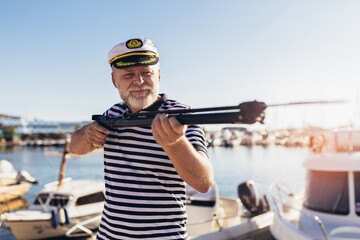 Obraz premium Mature man standing near the sea dressed in a sailor's shirt and hat holding a harpoon