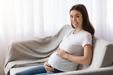 Portrait of beautiful young pregnant lady in casual clothes sitting on couch