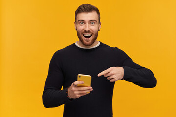 Bearded, happy looking man with brunette hair. Has piercing. Wearing black sweater. Holding and pointing finger at smartphone, copy space. Watching at the camera, isolated over yellow background