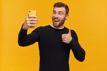 Bearded excited, happy looking man with brunette hair. Has piercing. Wearing black sweater. Making selfie and showing thumb up sign, approving gesture. Stand isolated over yellow background