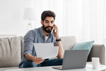 Arab freelancer man working remotely with smartphone, laptop and documents, sitting on sofa at home