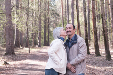 Fototapeta premium Elderly interracial couple in a spring forest park. A woman kissing a man on the cheek.