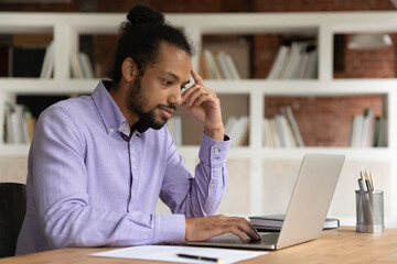 Focused thoughtful African American businessman looking at laptop screen, reading news in email, pensive young man employee freelancer pondering project strategy, solving problem, watching webinar