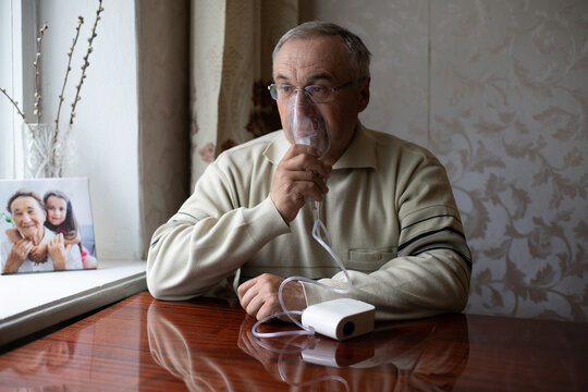 Portrait of elderly man with flu inhalation at home. The senior Caucasian man does inhalation with a nebulizer mask at home for coronavirus disease, sitting on couch at home alone. Health care concept