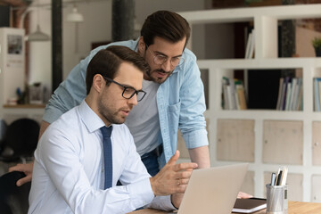 Confident mentor coach in glasses training intern, pointing at screen, helping to new worker with...