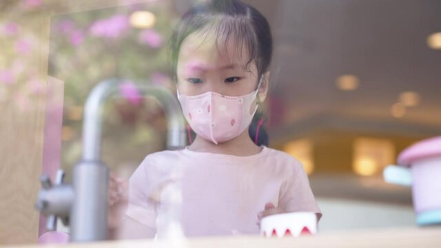 Medium close up scene behind the glass of Asian girl toddler wearing protective face mask playing kitchen toys for kid in the playground room. Social distancing during covid-19 pandemic.