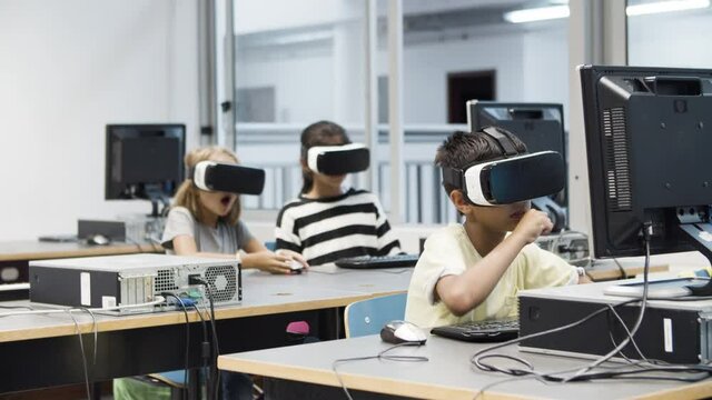 Multiethnic kids having fun using virtual reality headset. Funny blonde girl fooling and eating computer mouse. Children sitting and looking around. VR technology in education concept.