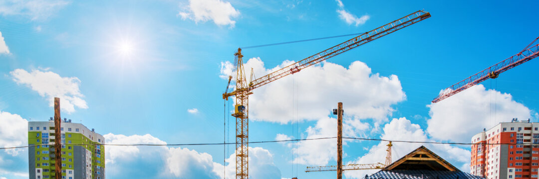 High construction cranes stands on building site against blue sky with sun.