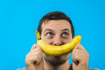 Giovane ragazzo si diverte creando un sorriso con una banana su sfondo blu - obrazy, fototapety, plakaty