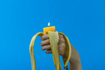 Mano di giovane ragazza tiene in pugno una banana sbucciata con all'interno una candela accesa dal basso su sfondo blu