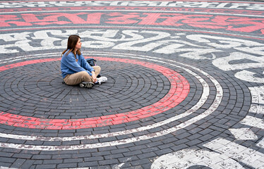 Young woman in blue sweatshirt alone sitting on street cobbled square in the form of circles and...