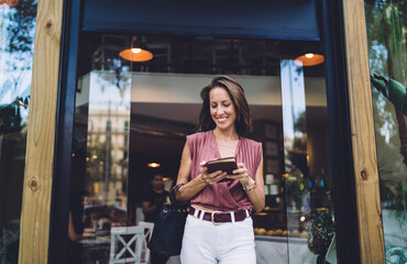 Prosperous caucasian female in trendy wear walking out of restaurant checking working plans from notebook, smiling business woman leaving own cafe enjoying entrepreneurship and lifestyle