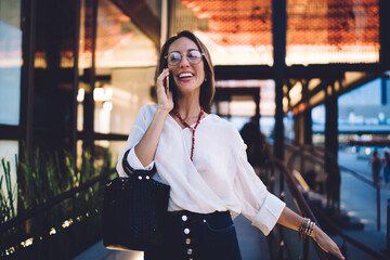 Cheerful Caucasian woman in optical spectacles laughing while receiving international call conversation on modern mobile phone, happy female user enjoying roaming internet connection for communicate