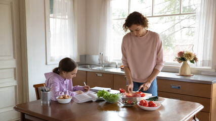 Caring young mother cooking healthy food while smart teenage kid daughter involved in doing homework, learning studying preparing assignment spending time together in kitchen, daily routine concept.