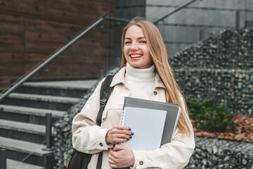 Happy girl student holding notebook folders in hands, smiling on college background