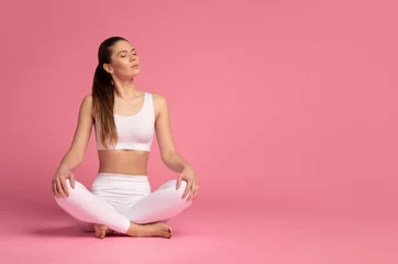 Fotobehang Young, beautiful woman in a white tracksuit practicing yoga meditation in the lotus position isolated on pink background, banner with place for text on the right © iso100production
