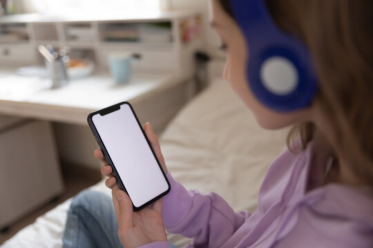 Close up young teenage girl in headphones holding smartphone in hands with mockup white screen, advertisement for children educational application or online game, modern technology addiction concept.