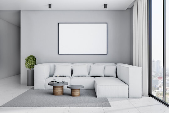 White poster with copyspace in black frame on light grey wall above big sofa in stylish spacious living room with plant in flower pot, coffee tables and city view from big window. 
