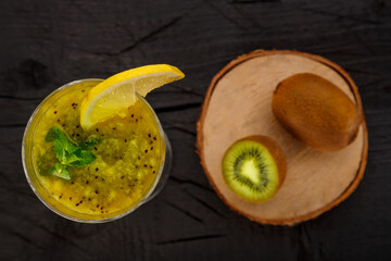 Kiwi smoothie garnished with mint and lemon in a glass on a black background near kiwi top view.