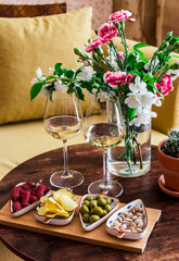 Two glasses of white wine and a variety of snacks olives, sausages, pistachios, chips on a wooden table with a bouquet of flowers in a cozy room