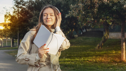 Young woman with closed eyes in white electronic headphones smiling and listening online audiobook in nature. Enjoying music or leisure podcast. Storytelling. Dreaming concept