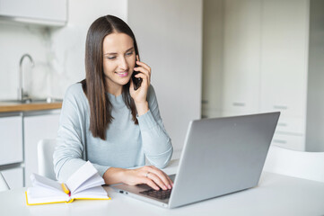 Charming young freelancer woman is talking on smartphone working with laptop from home, female employee has pleasant phone conversation working on the distance, connect with customers by mobile phone