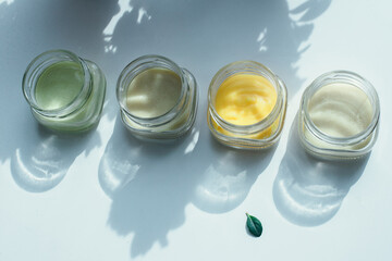 Top view of cosmetic cream in glass jars on white surface in a shadow of flowers with nice green leaf in front of them. Pastel colors, herbal cosmetics concept.