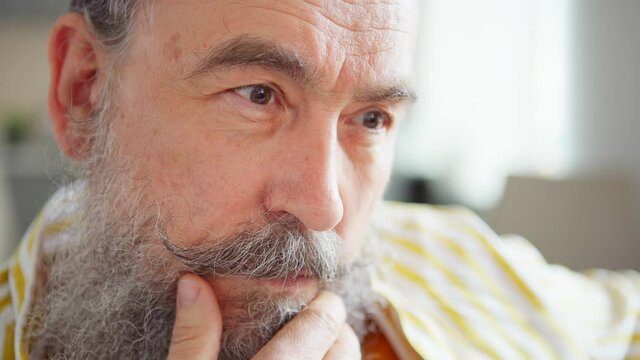 Closeup portrait of senior man touching his gray beard and mustache while thinking and looking at camera