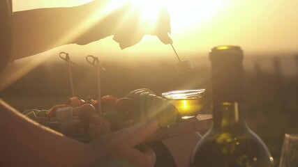 Against background of the bright sun, a woman's hand lowers a skewer of cheese (canapes) into plate of honey. a board with sliced fruit on the background of sunrise and sunset, Romance, bottle wine - Powered by Adobe