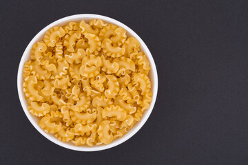 White glass bowl of raw pasta. Isolated on a dark grey background. Top view close up