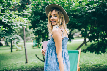 Obraz premium Half length portrait of joyful female student with education textbook posing during daytime in city park, happy Caucasian hipster girl in stylish hat holding personal planner and smiling at camera