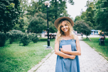 Half length portrait of joyful female student with education textbook posing during daytime in city park, happy Caucasian hipster girl in stylish hat holding personal planner and smiling at camera