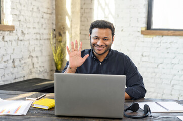 Hindu male office employee holding video meeting sitting at the desk in loft office, cheerful businessman in smart casual shirt greeting colleagues, has virtual conference