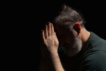 Bearded man holding hands in prayer in the sun, face hidden in shadow,  surrounded with darkness