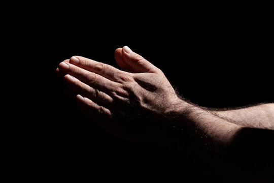 Male hands folded in prayer in the sunlight with harsh shadows, surrounded with darkness 