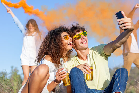 Beautiful young man and woman hold smartphone, doing selfie - Happy friends having fun in the park with multicolored smoke bombs smoke. Young students celebrating spring break together. Holi festival.