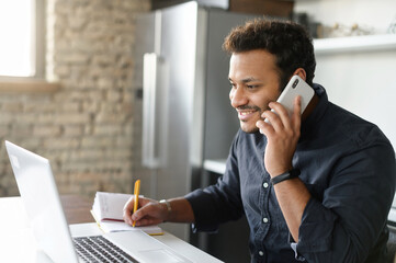 Happy indian guy in smart casual shirt talking on the smartphone and using laptop for remote work from home, mixed-race male entrepreneur takes notes during phone conversation, his business going well