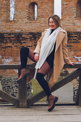 woman sitting on a bench, Beautiful happy smiling young woman sits on fence in a beige coat, white wool cashmere scarf and brown boots, street style photography. Film noise, soft focus